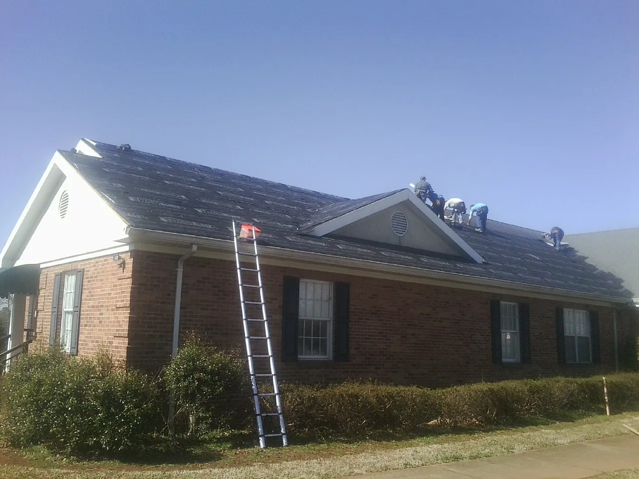 Crew tearing shingles off a brick ranch roof with a ladder set — Metal Roof Repair work by our crew in Chocowinity