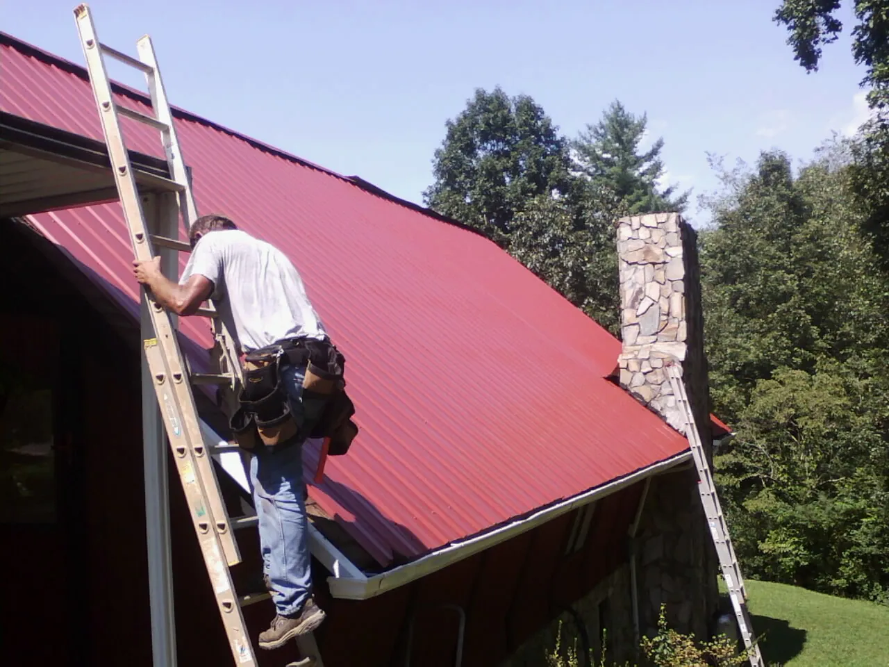 Roofer on a ladder beside a red metal roof and stone chimney — Soffit & Fascia Repair by Roofing Brothers in Chocowinity